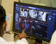 Aluno do curso de Radiologia analisa exames de imagem em um monitor de alta resolução. A tela exibe múltiplas imagens médicas, incluindo cortes axiais e reconstruções 3D de órgãos internos. Ambiente moderno, com materiais de escritório ao redor, reforçando a prática acadêmica em diagnóstico por imagem.