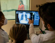 Grupo de estudantes de Radiologia observa e discute uma radiografia de tórax exibida em monitor, enquanto uma paciente aguarda posicionada na sala ao fundo. A equipe usa jalecos brancos e máscaras, destacando o ambiente seguro e profissional do laboratório universitário dedicado à análise de exames radiológicos.