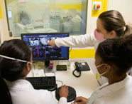 Três estudantes de Radiologia, usando jalecos e máscaras, analisam exames de imagem em um computador dentro do laboratório da faculdade. Uma delas aponta para detalhes na tela, enquanto ao fundo é possível ver o aparelho de tomografia através de uma janela, ilustrando a integração entre teoria e prática no ensino superior.