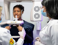 Em laboratório de Radiologia, estudante realiza exame de imagem odontológica utilizando equipamento panorâmico. A pessoa examinada veste avental de proteção, enquanto duas alunas acompanham o procedimento, todas de jaleco branco e máscara, evidenciando a formação prática em diagnóstico por imagem da instituição.