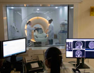 Ambiente de ressonância magnética em instituição de ensino, com duas estudantes de Radiologia posicionando paciente no aparelho circular iluminado. Na sala de controle, outra aluna monitora as imagens cerebrais em dois monitores, demonstrando a rotina prática do curso de Radiologia no diagnóstico avançado.