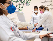 Equipe de estudantes de Radiologia prepara paciente para exame de densitometria óssea em laboratório da instituição. Paciente deitado sobre a mesa do equipamento, enquanto alunas ajustam sua posição. Em primeiro plano, profissional monitora o exame no computador, todos usando jaleco e máscara.