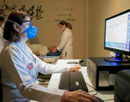 Aluna do curso de Radiologia opera computador durante exame de densitometria óssea, enquanto colegas preparam paciente deitado no aparelho. Todos usam jaleco branco e máscaras, reforçando o ambiente clínico e seguro da instituição de ensino voltada à formação em diagnóstico por imagem.