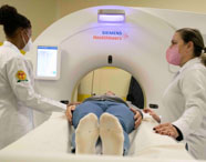 Paciente deitado em mesa de tomógrafo Siemens Healthineers, sendo preparado por duas estudantes de Radiologia em jaleco branco. Vista frontal do equipamento, destacando o cuidado dos alunos durante o exame prático em ambiente moderno de ensino superior na área de diagnóstico por imagem.