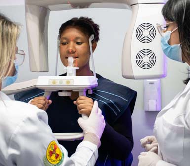 Alunos em laboratório de radiologia, com estudante usando avental de proteção sendo posicionado para exame de imagem odontológica por duas profissionais de jaleco branco e máscara. Equipamento moderno de radiografia panorâmica envolve a cabeça do paciente. Ambiente clínico, típico de curso técnico ou superior em Radiologia na instituição de Prudente.