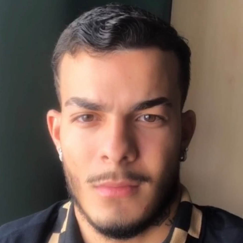 Kauã, aluno de Educação Física - Bacharelado, posando em um ambiente interno com um fundo neutro. Ele tem barba e bigode, e usa brincos de argola, com uma expressão séria.