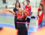 Estudante de Educação Física realiza atividade com bambolê rosa em quadra esportiva coberta, usando uniforme preto e vermelho. Ao fundo, colegas observam e participam, alguns vestindo coletes vermelhos ou roupas casuais. O ambiente é colorido, com piso azul e verde, típico de ginásio universitário.