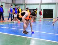 Aluno de Educação Física, com colete laranja, participa de dinâmica colocando cone azul no chão de quadra poliesportiva. Outros estudantes, com coletes azuis e amarelos, aguardam ao fundo. O espaço é amplo, com piso marcado para esportes e redes, evidenciando estrutura de ginásio acadêmico.