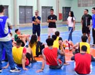 Grupo de estudantes de Educação Física da Unoeste, campus Presidente Prudente, sentados no chão da quadra, ouvindo orientações de colegas em pé. Coletes coloridos identificam equipes. O ginásio possui piso azul e laranja, redes de gol e paredes claras, caracterizando espaço de práticas esportivas.