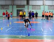 Gincana de Educação Física em quadra coberta, com estudante agachado pegando bambolê rosa entre cones coloridos. Ao fundo, grupos de alunos com coletes de diferentes cores aguardam sua vez. Ambiente vibrante, com marcações esportivas e estrutura típica de ginásio universitário.
