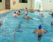 Aula prática de natação ou polo aquático com estudantes de Educação Física em piscina coberta. Alunos usam toucas coloridas e disputam bola na água, enquanto pequenas traves flutuam. O ambiente é iluminado, com bordas de azulejo claro e materiais esportivos ao fundo.