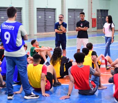 Grupo de estudantes do curso de Educação Física da Unoeste, campus Presidente Prudente, participa de atividade prática em quadra esportiva coberta. Alunos vestem coletes coloridos e estão sentados no chão, atentos a orientações de colegas em pé. Ao fundo, redes de gol e paredes claras compõem o ambiente esportivo e acadêmico.