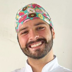 Bruno, aluno do curso de odontologia, vestindo jaleco branco e um gorro colorido com estampas divertidas. Ele está em um ambiente interno com fundo claro.
