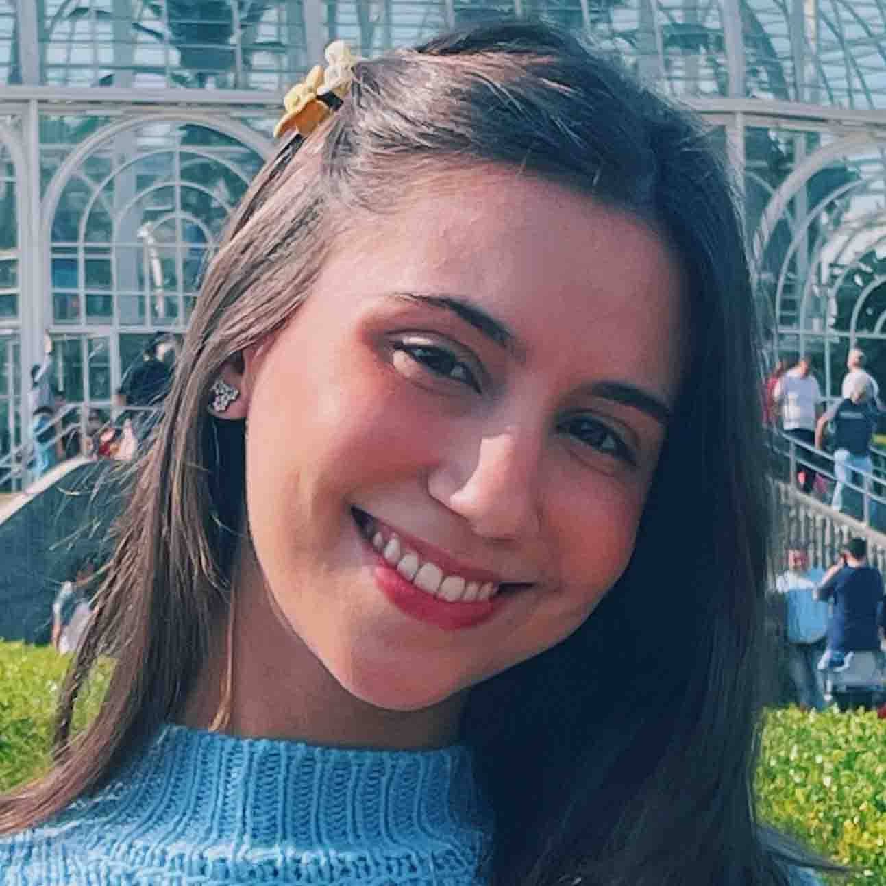 Maria Clara, aluna do curso de odontologia, em um ambiente externo com uma estrutura de vidro ao fundo. Ela veste uma blusa azul e usa um acessório no cabelo, compondo um visual descontraído.