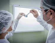 Estudantes de Odontologia analisam uma radiografia odontológica em frente a um negatoscópio. Ambos usam jaleco branco, touca e luvas descartáveis. Um deles segura a radiografia com pinça metálica enquanto o outro observa atentamente, evidenciando o ambiente clínico-acadêmico da instituição.