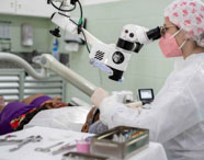 Estudante de Odontologia realiza atendimento em consultório equipado, utilizando microscópio odontológico branco articulado. A aluna veste jaleco, touca estampada e luvas, enquanto paciente repousa na cadeira. O cenário é moderno, com instrumentos organizados e ambiente limpo, característico de laboratório de habilidades clínicas.