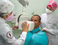 Duas estudantes de Odontologia, com jaleco branco e toucas, posicionam aparelho de raio-x odontológico próximo à cabeça de um paciente sentado, que veste avental protetor azul. Ambiente de clínica universitária, parede verde-clara e equipamentos modernos, destacando a prática supervisionada na formação acadêmica.