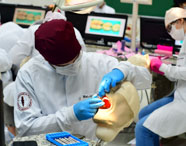 Estudante de Odontologia pratica procedimentos em manequim odontológico, vestindo jaleco branco, touca vinho e luvas azuis. Ao fundo, outros alunos trabalham em manequins semelhantes, com monitores exibindo imagens de dentes. Ambiente de laboratório, organizado e tecnológico, voltado para treinamento prático.