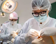 Alunos do curso de Odontologia realizam atividades práticas em manequins odontológicos, sob luzes cirúrgicas. Todos usam jaleco branco, touca e luvas, concentrados nos procedimentos. O ambiente é amplo, com várias estações de trabalho e iluminação adequada, reforçando o ensino técnico em laboratório universitário.