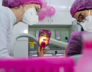 Duas estudantes de Odontologia, com jaleco branco e máscaras, atendem paciente infantil que segura espelho rosa. O consultório tem decoração com balões cor-de-rosa e armários brancos ao fundo. Cena demonstra acolhimento e atenção durante atendimento clínico em ambiente universitário.