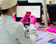 Estudante de Odontologia, de costas, com jaleco branco e touca bege, manipula ponteiras odontológicas dentro de estojo rosa. Usa luvas rosas e está diante de bancada com instrumentos, tubos e computador exibindo ilustrações anatômicas. Ambiente de laboratório prático, organizado e colorido.