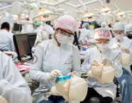 Sala ampla de laboratório odontológico com vários estudantes praticando em manequins de cabeça humana. Todos usam jaleco branco, touca e luvas, sentados em bancadas equipadas com computadores e instrumentos. Ambiente iluminado, propício ao desenvolvimento de habilidades técnicas no curso de Odontologia.