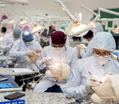 Estudantes do curso de Odontologia da Unoeste em Presidente Prudente realizam prática em laboratório, utilizando manequins odontológicos para simulação de procedimentos. Todos usam jaleco branco, toucas e luvas, sentados em bancadas equipadas com refletores individuais, instrumentos e materiais odontológicos.