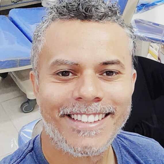 Diego Silva dos Santos, aluno de Engenharia Elétrica, posando em um ambiente interno com equipamentos médicos ao fundo. Ele tem cabelos grisalhos e barba, com um sorriso largo.