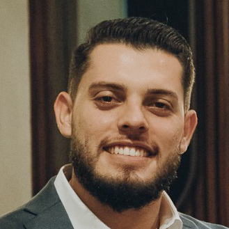 Henrique, egresso de Engenharia Elétrica, posando em um ambiente interno com um fundo neutro. Ele tem barba e um sorriso largo.