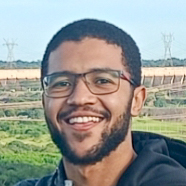 Sebastião, aluno de Engenharia Elétrica, posando em um ambiente externo com uma paisagem industrial ao fundo. Ele usa óculos e tem barba, com um sorriso largo.