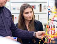 Aluna observa atentamente enquanto professor manipula fios amarelos e vermelhos conectados a painel didático de instrumentação elétrica. O equipamento possui botões, luzes e tomadas, típico de laboratórios do curso de Engenharia Elétrica, ambiente repleto de painéis ao fundo.