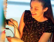 Estudante de Engenharia Elétrica opera painel de instrumentação, conectando cabos azuis e pretos em entradas específicas. Veste blusa preta estampada e está envolvida em experimento prático, evidenciando a aplicação de conceitos elétricos em laboratório universitário.