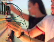Dois estudantes realizam ajustes em painel de instrumentação elétrica, conectando cabos azuis e verdes em diferentes pontos. O painel exibe medidores analógicos, botões e luzes indicadoras, compondo cenário típico de laboratório de Engenharia Elétrica.