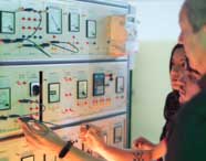 Close em miliamperímetro DC instalado em painel de instrumentação elétrica, cercado por botões, mostradores digitais e conexões coloridas. Equipamento utilizado por estudantes de Engenharia Elétrica para medições precisas em experimentos laboratoriais