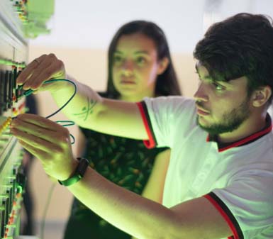 Estudante do curso de Engenharia Elétrica conecta fios em painel didático repleto de botões, tomadas e luzes indicadoras. O ambiente é iluminado por luz esverdeada, destacando o trabalho prático em laboratório da instituição, com foco em circuitos elétricos e automação.