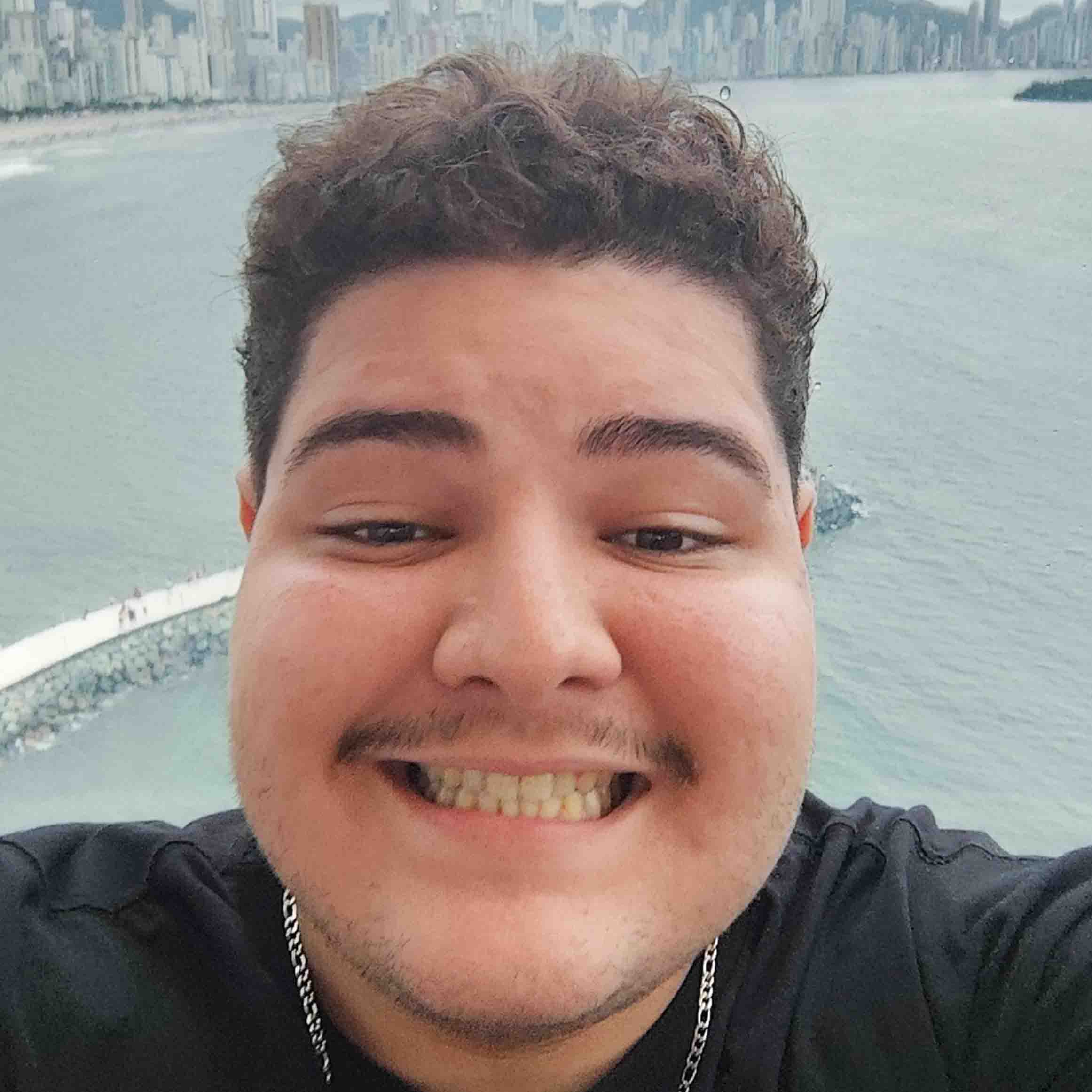Andrei, aluno de Pedagogia, posando em um ambiente externo com uma cidade e o mar ao fundo. Ele tem bigode e um sorriso largo.