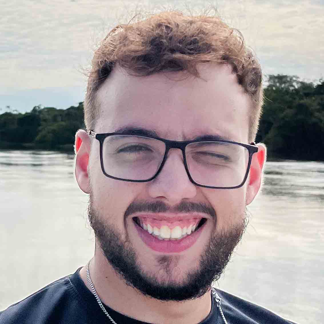 Natan, aluno de Pedagogia, posando em um ambiente externo com um rio e folhagens ao fundo. Ele usa óculos de armação preta e tem barba, com um sorriso largo.