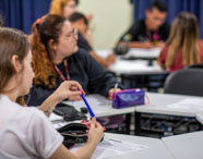 Estudantes do curso de Pedagogia acompanham aula em sala, sentados em mesas brancas com folhas e estojos coloridos. Uma aluna segura uma caneta azul, enquanto outros escrevem ou prestam atenção. O ambiente é acadêmico, com cadeiras cinza e atmosfera de concentração durante atividade teórica.