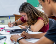Alunos de Pedagogia realizam atividades escritas em sala de aula. Uma estudante de cabelos longos escreve no caderno, rodeada por materiais como estojo preto, celular rosa e canetas. O quadro verde ao fundo e a disposição das mesas evidenciam o ambiente universitário da Unoeste.