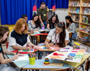 Turma de Pedagogia participa de atividade prática em brinquedoteca, sentados em mesas coloridas. Estão desenhando e pintando cartazes, com materiais pedagógicos como lápis de cor, potes de tinta e jogos educativos. Ao fundo, prateleiras com livros e decoração lúdica criam clima acolhedor e criativo.
