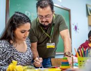 Professor orienta estudante de Pedagogia durante atividade lúdica na sala de aula da Unoeste. A aluna trabalha com materiais coloridos sobre a mesa, incluindo lápis, papéis e brinquedos pedagógicos. O quadro verde ao fundo reforça o contexto acadêmico e de formação docente.