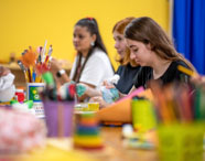 Estudantes de Pedagogia participam de atividade prática de pintura em sala decorada com paredes amarelas e cortina azul. Mesas estão repletas de pincéis, tintas e objetos pedagógicos. Alunas interagem, trocando ideias e materiais, demonstrando envolvimento com práticas educativas criativas.