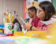 Alunos do curso de Pedagogia desenvolvem atividades manuais, recortando papel e utilizando pincéis e tintas guache. As mesas estão cheias de materiais escolares, como potes de tinta, tesouras e recipientes para pincéis. O ambiente remete à brinquedoteca universitária, estimulando criatividade e didática.