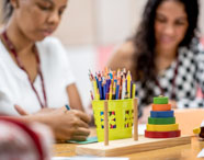 Duas estudantes de Pedagogia trabalham em atividades pedagógicas, desenhando e escrevendo em folhas coloridas. Na mesa, há um recipiente amarelo com lápis de cor e um brinquedo educativo de argolas coloridas empilháveis, reforçando o foco em práticas lúdicas para formação docente.