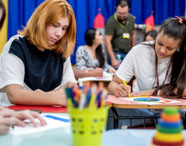 Estudantes de Pedagogia realizam atividades artísticas em brinquedoteca, sentados em mesas coloridas. Utilizam lápis de cor, papéis e outros materiais didáticos. O ambiente é vibrante, com cortina azul e parede amarela, promovendo integração entre teoria e prática no ensino superior.