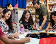 Grupo de alunos de Pedagogia recebe orientação de professor durante aula prática em brinquedoteca. Sentados em círculo, utilizam materiais pedagógicos como lápis de cor, cartolinas e jogos. Ao fundo, estantes com livros e decoração lúdica, ambiente típico de formação educacional na Unoeste.