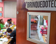 Porta de madeira aberta revela sala de brinquedoteca, onde estudantes de Pedagogia participam de atividade prática em grupo. Mesas repletas de tintas, pincéis e materiais coloridos. Ambiente lúdico, com paredes rosas e quadros decorativos, propício ao desenvolvimento de práticas pedagógicas.