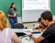 Professora ministra aula para turma de Pedagogia na Unoeste, em sala com quadro verde e projetor exibindo conteúdo didático. Alunos sentados nas primeiras fileiras escrevem em cadernos, atentos à explicação. Ambiente moderno, organizado e voltado para formação de futuros educadores.