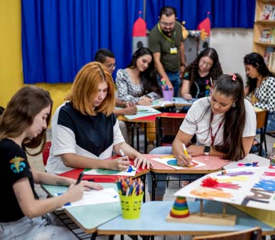 Sala de aula colorida com estudantes do curso de Pedagogia da UNOESTE Prudente sentados em mesas redondas, realizando atividades artísticas com papel, lápis de cor e canetinhas. Ao fundo, há estante de livros, cortina azul e decoração lúdica com castelos, criando ambiente acolhedor e criativo para formação docente.