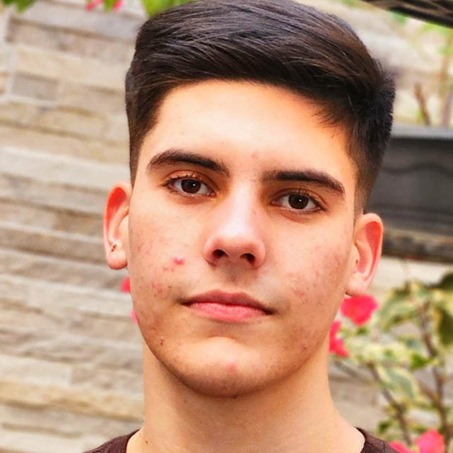 Bruno, aluno de Engenharia Mecânica, posando em um ambiente externo com uma parede de tijolos e plantas ao fundo. Ele tem cabelos escuros e uma expressão séria.